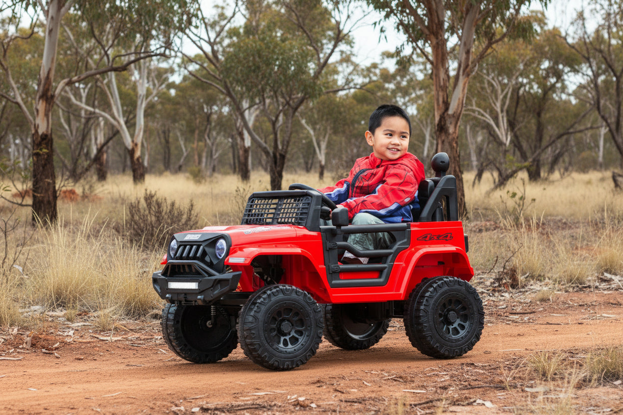 Child sitting in a red toy truck outdoors with greenery in the background